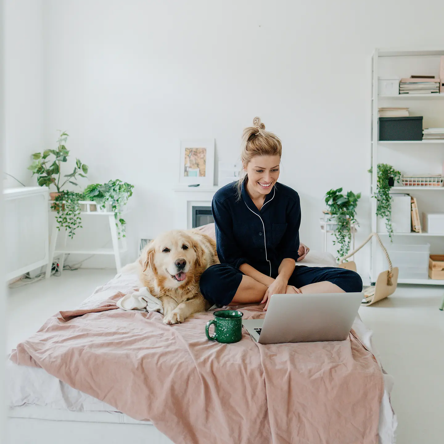 Eine Frau sitzt im Schlafanzug mit Notebook und Hund auf dem Bett