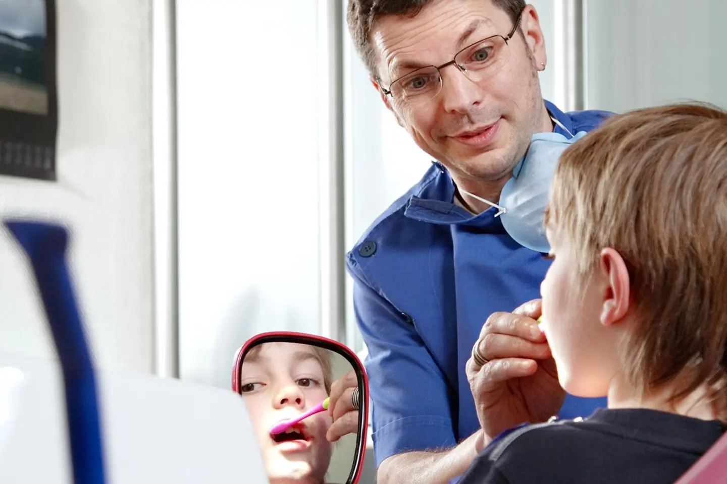 Ein Junge sitzt beim Zahnarzt auf dem Stuhl und schaut in einen Spiegel. Der Arzt zeigt und erklärt ihm etwas.