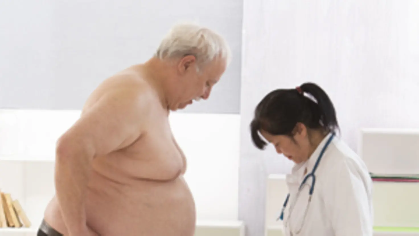 Ein übergewichtiger Mann hat einen Fuß auf einer Waage, eine Arzt-Schwester steht neben ihm und erklärt ihm etwas.