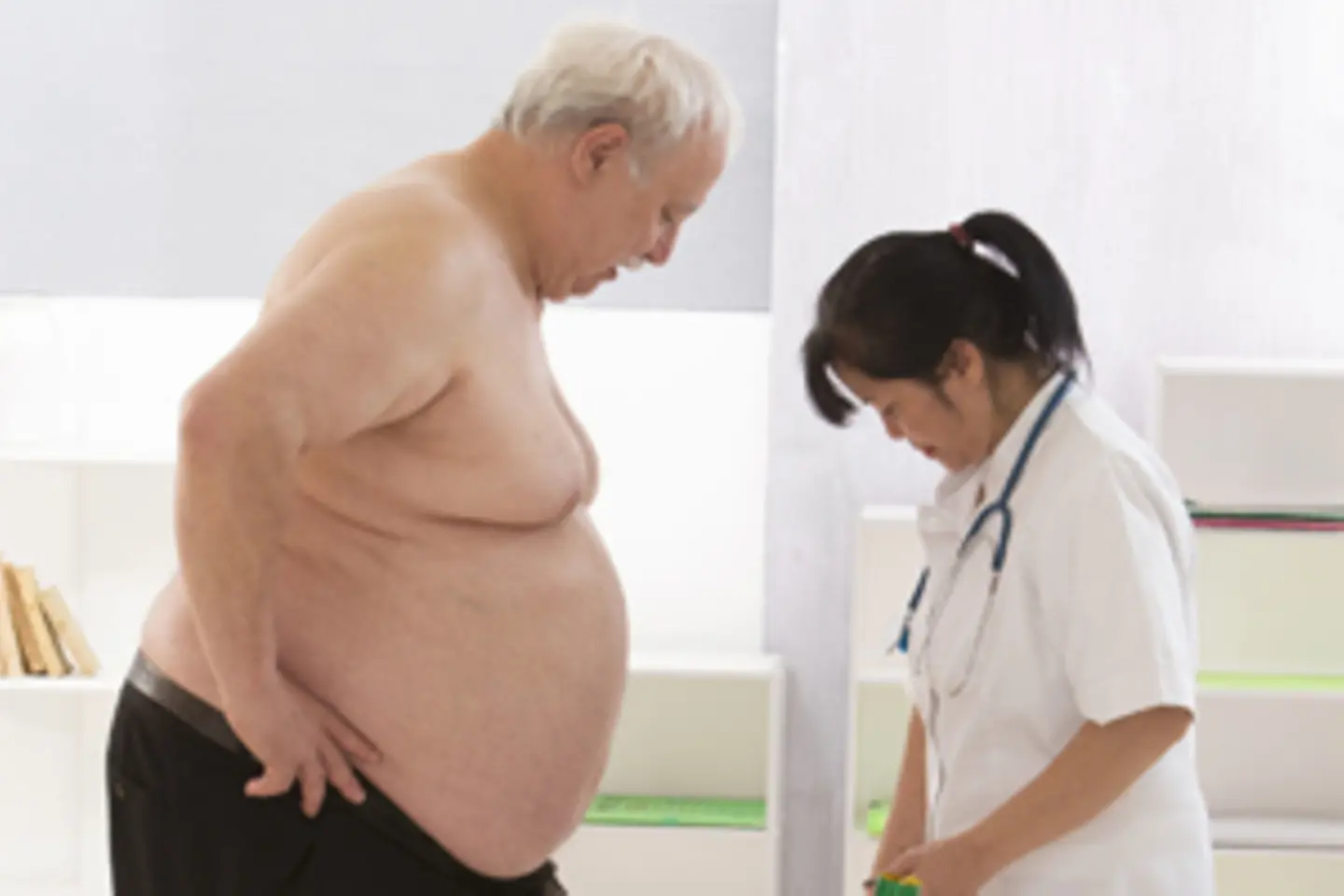 Ein übergewichtiger Mann hat einen Fuß auf einer Waage, eine Arzt-Schwester steht neben ihm und erklärt ihm etwas.