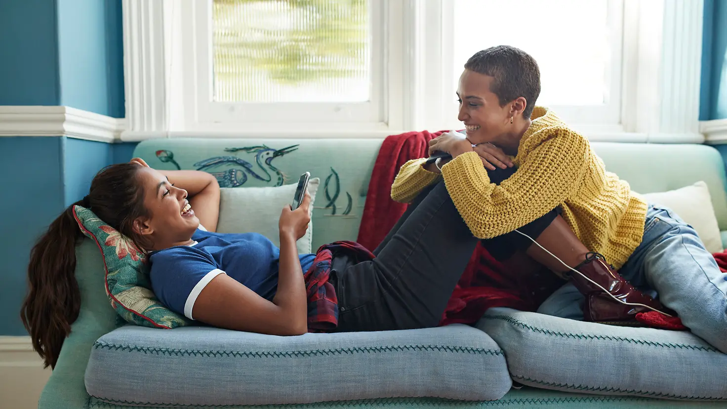 Zwei junge Frauen liegen mit einem Handy auf einem Sofa und lachen sich an