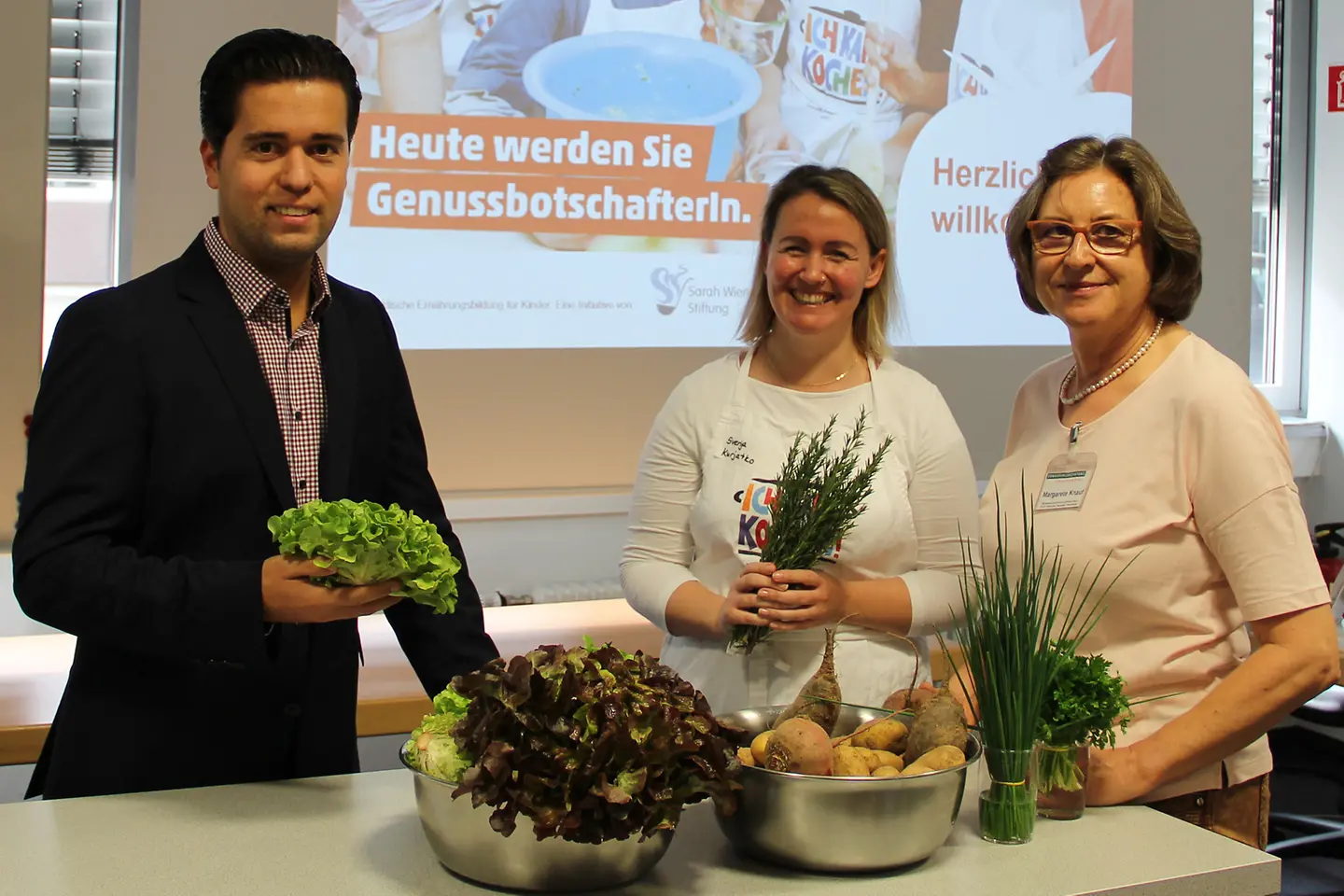 Dominik Bergemann, Fortbildungsleiterin Kurjatko und Maragarete Knauf vor einer Wand mit Gemüse in der Hand.