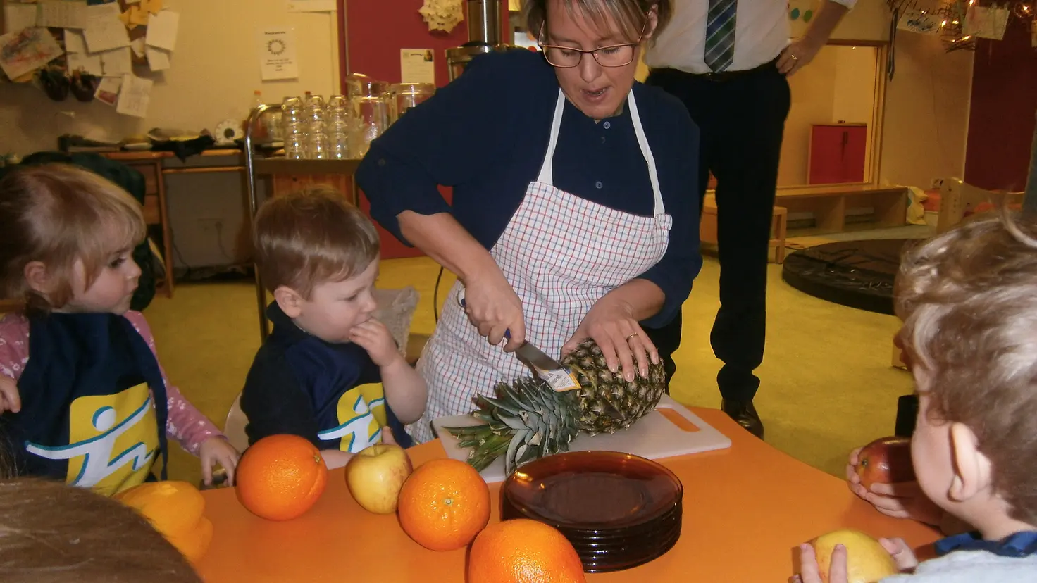 KITA-Mitarbeiterin zeigt Kindern, wie man eine Ananas schneidet.