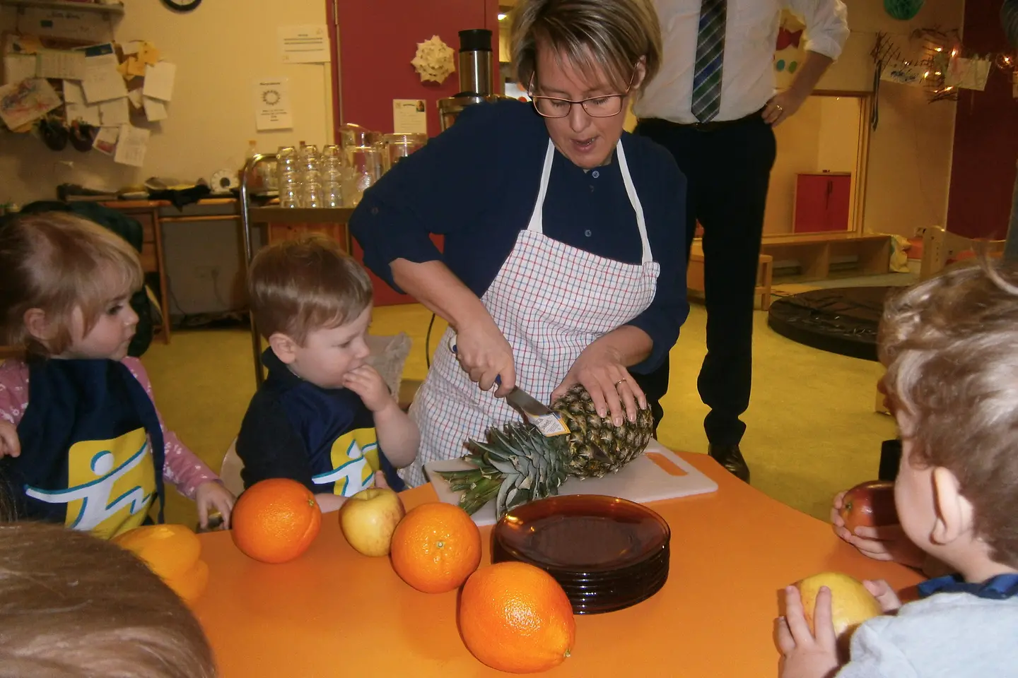 KITA-Mitarbeiterin zeigt Kindern, wie man eine Ananas schneidet.