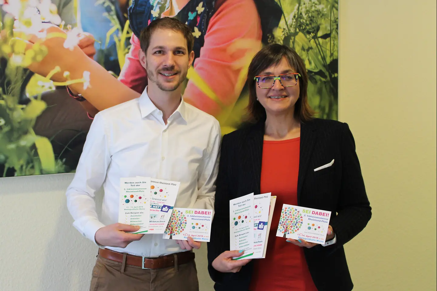 Dunja Kleis und Johannes Schweizer mit Broschüren und Flyern zur Inklusionsmesse vor einer Wand.
