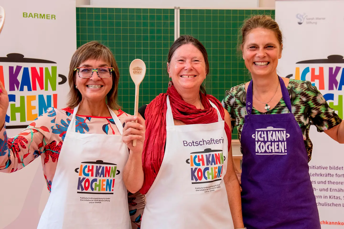 Dunja Kleis, Ulrike Höfken und Sarah Wiener heben je einen Kochlöffel in die Höhe stehend vor einer Schultafel.