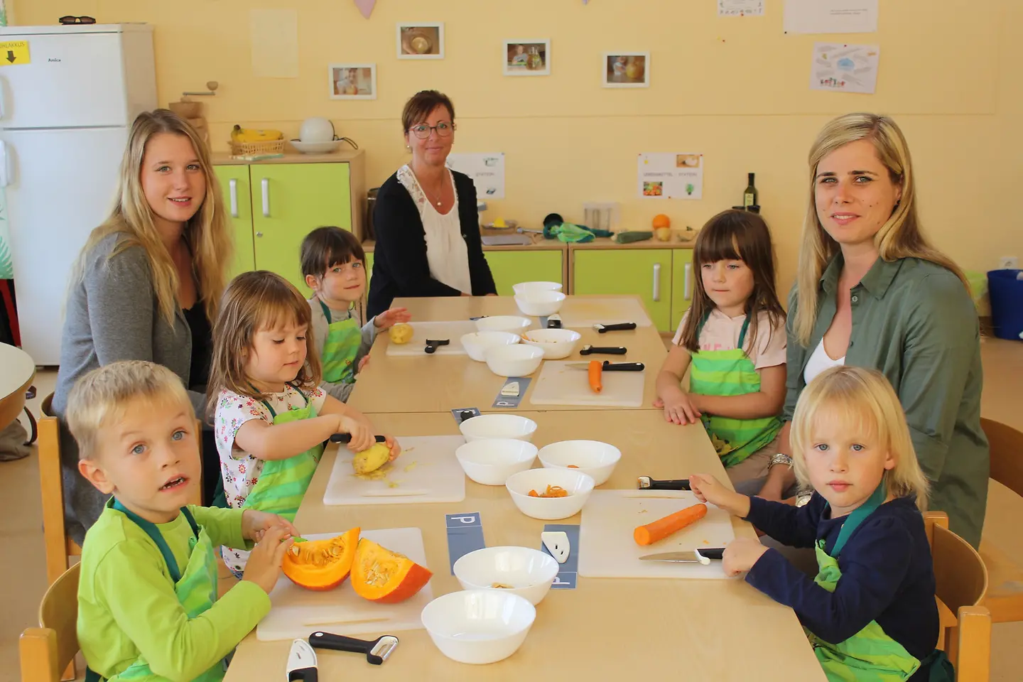 Erzieherinnen und BARMER Vertreterin sitzen mit Kindern an einem Tisch und schnippeln Gemüse.