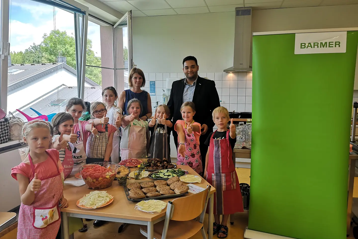 Kinder in einer Küche strecken den Daumen nach oben zusammen mit ihrer Lehrerin und einem BARMER Mitarbeiter.