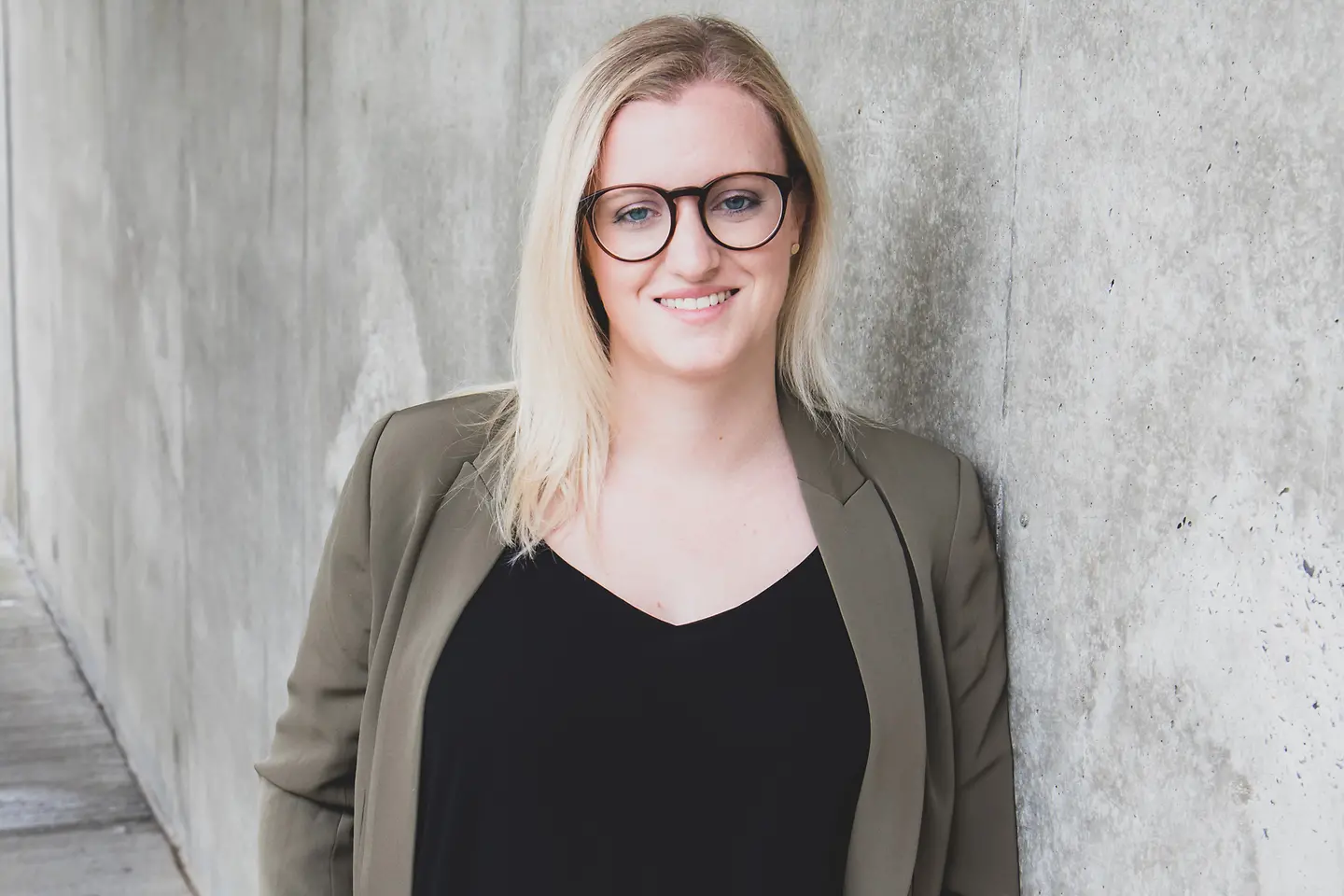 Sarah Klein: eine junge Frau mit langen blonden Haaren und schwarzer Brille lehnt an einer Wand und lächelt in die Kamera