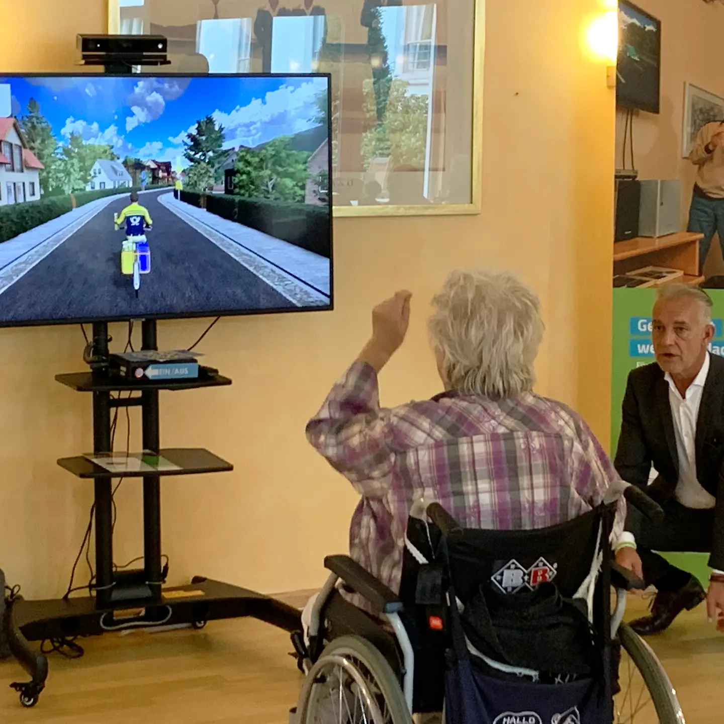 Eine Seniorin sitzt im Rollstuhl von einem Bildschirm auf dem ein Postbote auf dem Fahrrad fährt. 