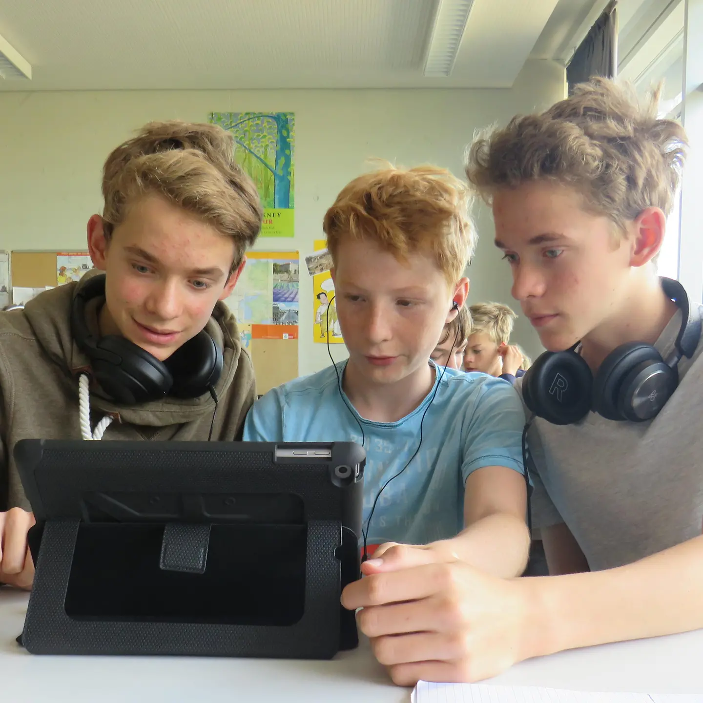 Drei Jungen im Alter von 14 Jahren sitzen im Klassenzimmer vor einem Laptop und haben Kopfhörer um den Hals.
