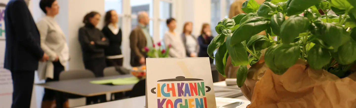 Ein Foto mit Obst und Basilikum im Vordergrund und Teilnehmern im Hintergrund auf einer Fortbildung "Ich kann kochen".