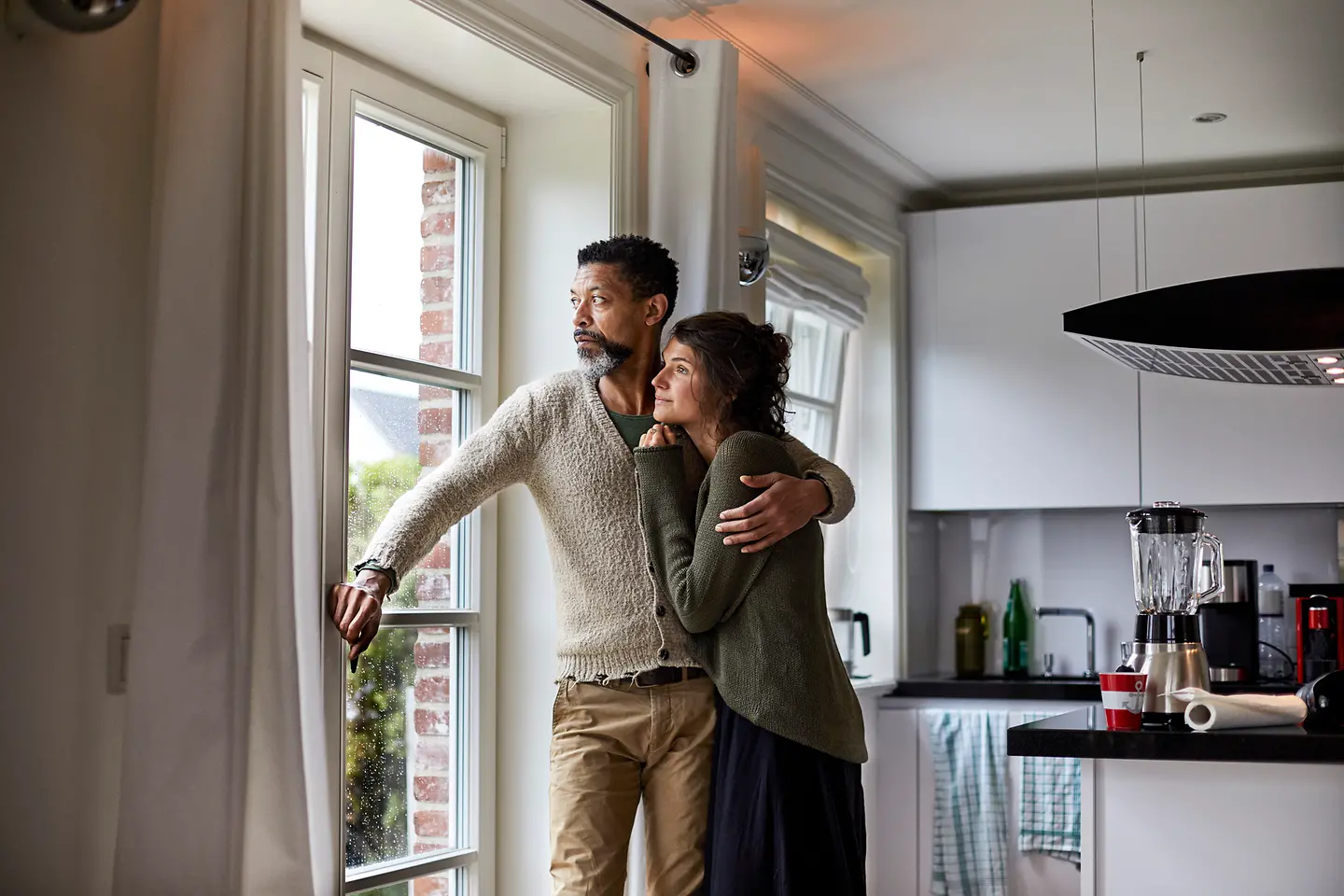 Ernster Mann umarmt eine Frau, sie schauen aus einem Fenster
