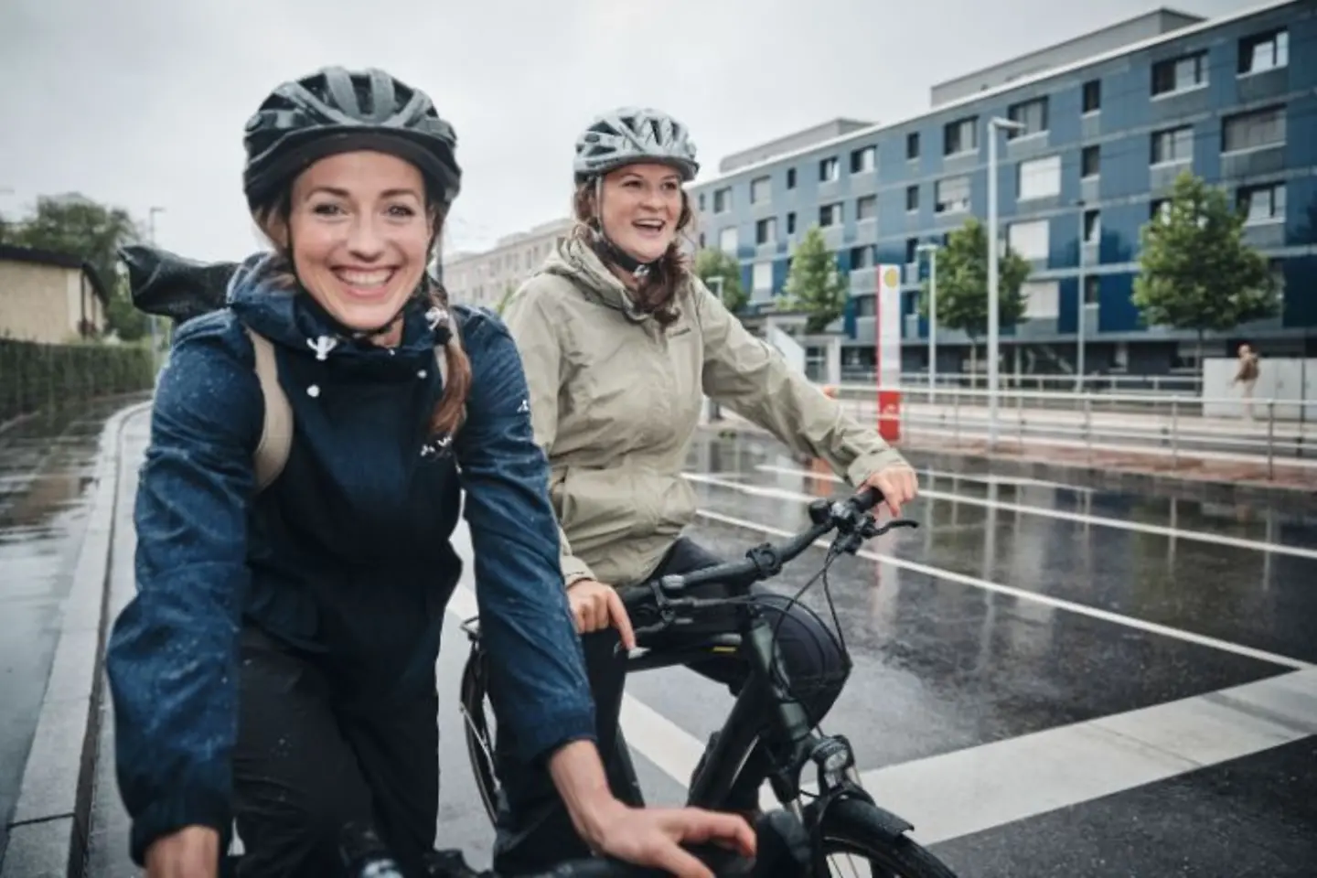 Zwei Frauen stehen bei Regen mit ihrem Fahrrad an einer Kreuzung und lachen in die Kamera.