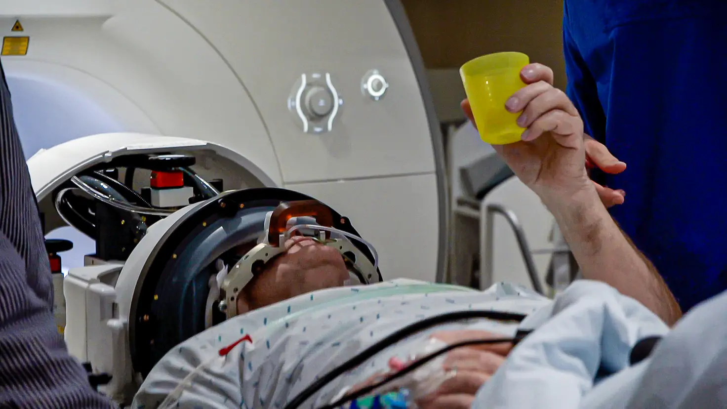 Ein Patient liegt mit einem Tremor-Helm in einem Magnetresonanztomographen und hält einen Plastikbecher in der Hand