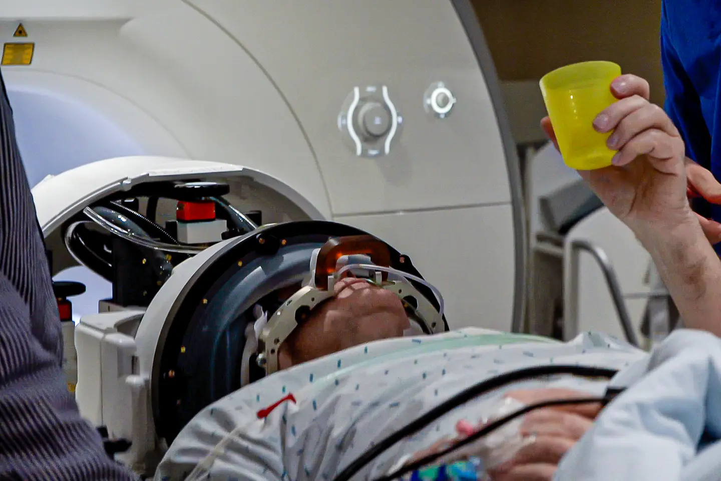 Ein Patient liegt mit einem Tremor-Helm in einem Magnetresonanztomographen und hält einen Plastikbecher in der Hand