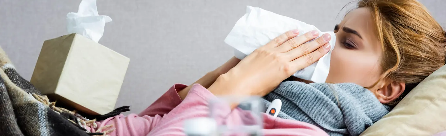 Eine Frau liegt mit Grippe im Bett und schnäuzt sich die Nase.
