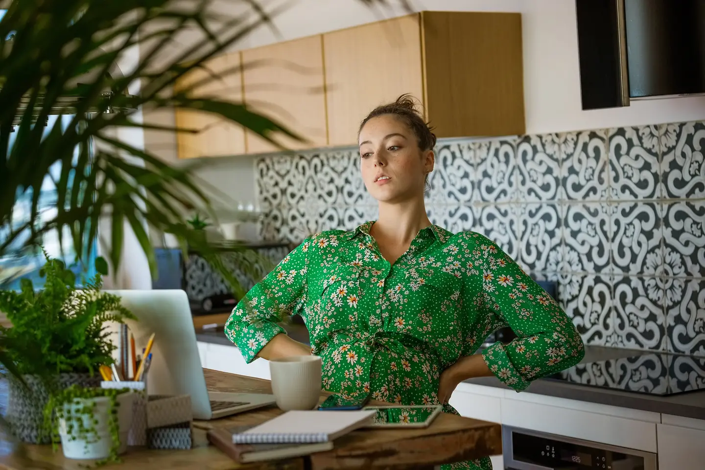Eine junge schwangere Frau im grünen Kleid sitzt an einem Tisch und schaut auf ein Notebook-Screen.