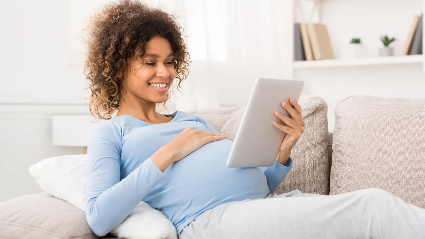 Eine schwangere Frau liegt auf dem Sofa und schaut auf ein Tablet, das sie in ihrer Hand hält.