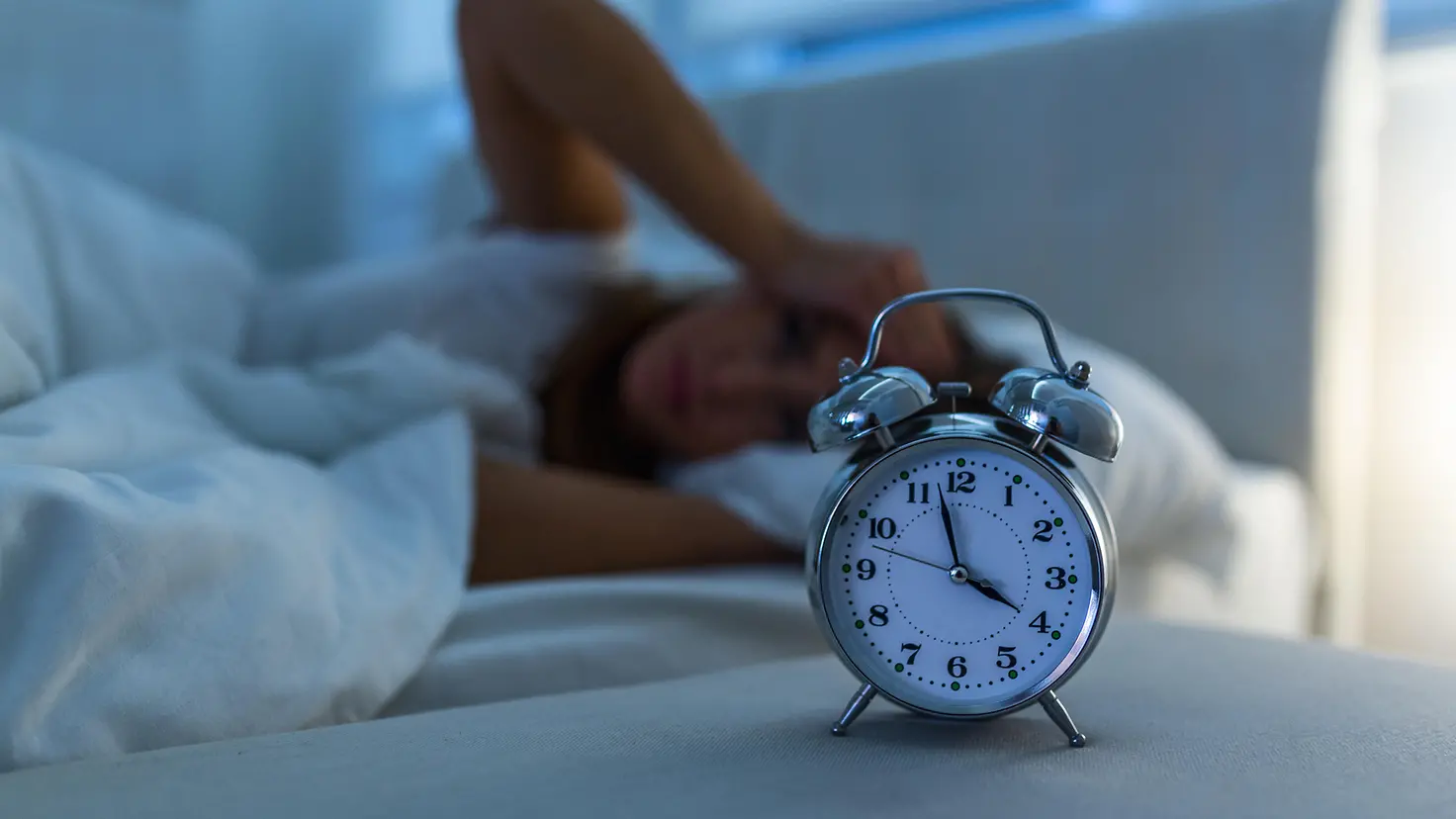 Das Bild zeigt eine Frau, die im Bett liegt und nicht schlafen kann. Vor ihr steht ein Wecker, der anzeigt, dass es 4 Uhr ist.