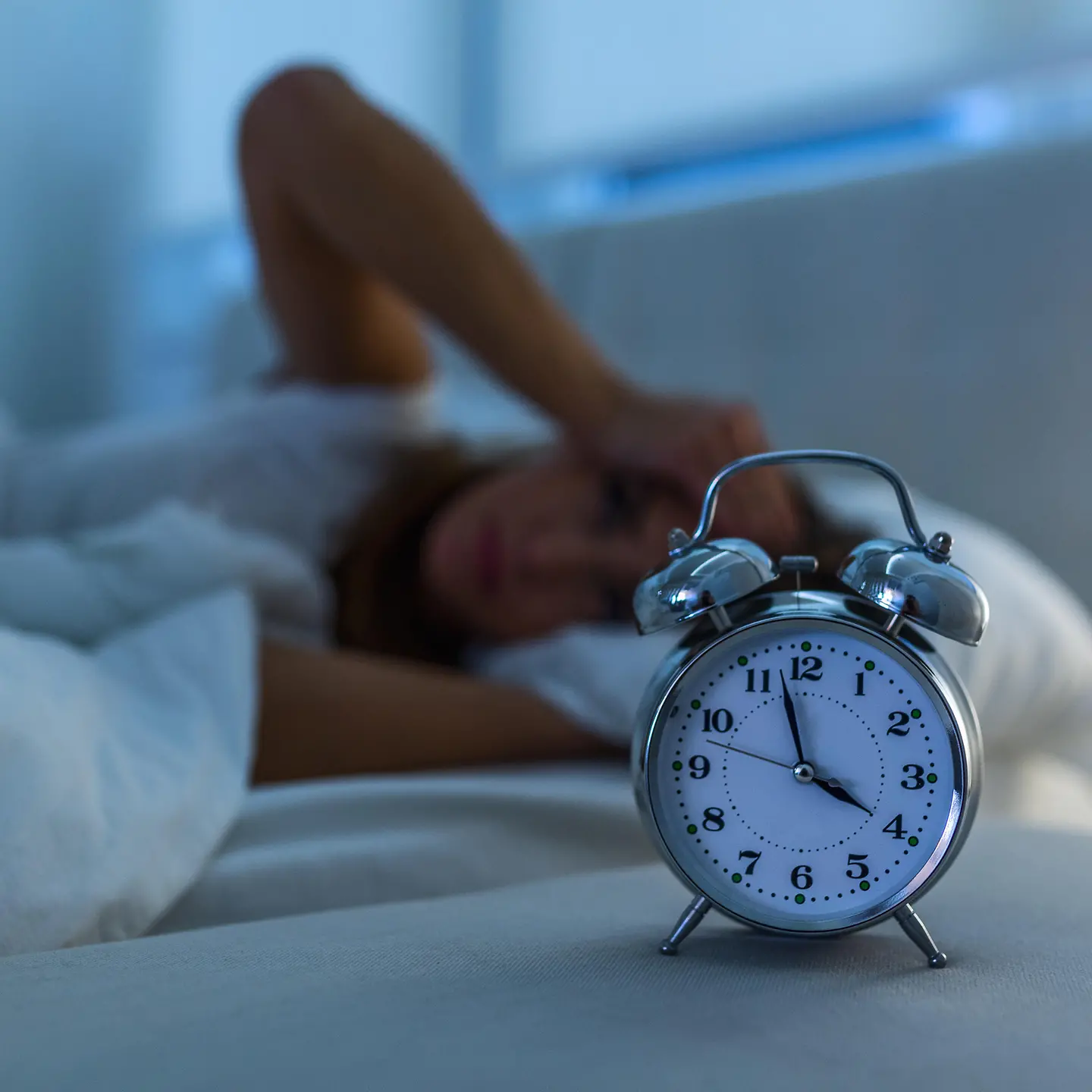Das Bild zeigt eine Frau, die im Bett liegt und nicht schlafen kann. Vor ihr steht ein Wecker, der anzeigt, dass es 4 Uhr ist.
