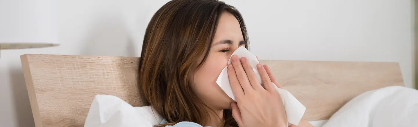 Das Bild zeigt eine Frau, die mit einer Erkältung im Bett liegt und sich die Nase putzt.