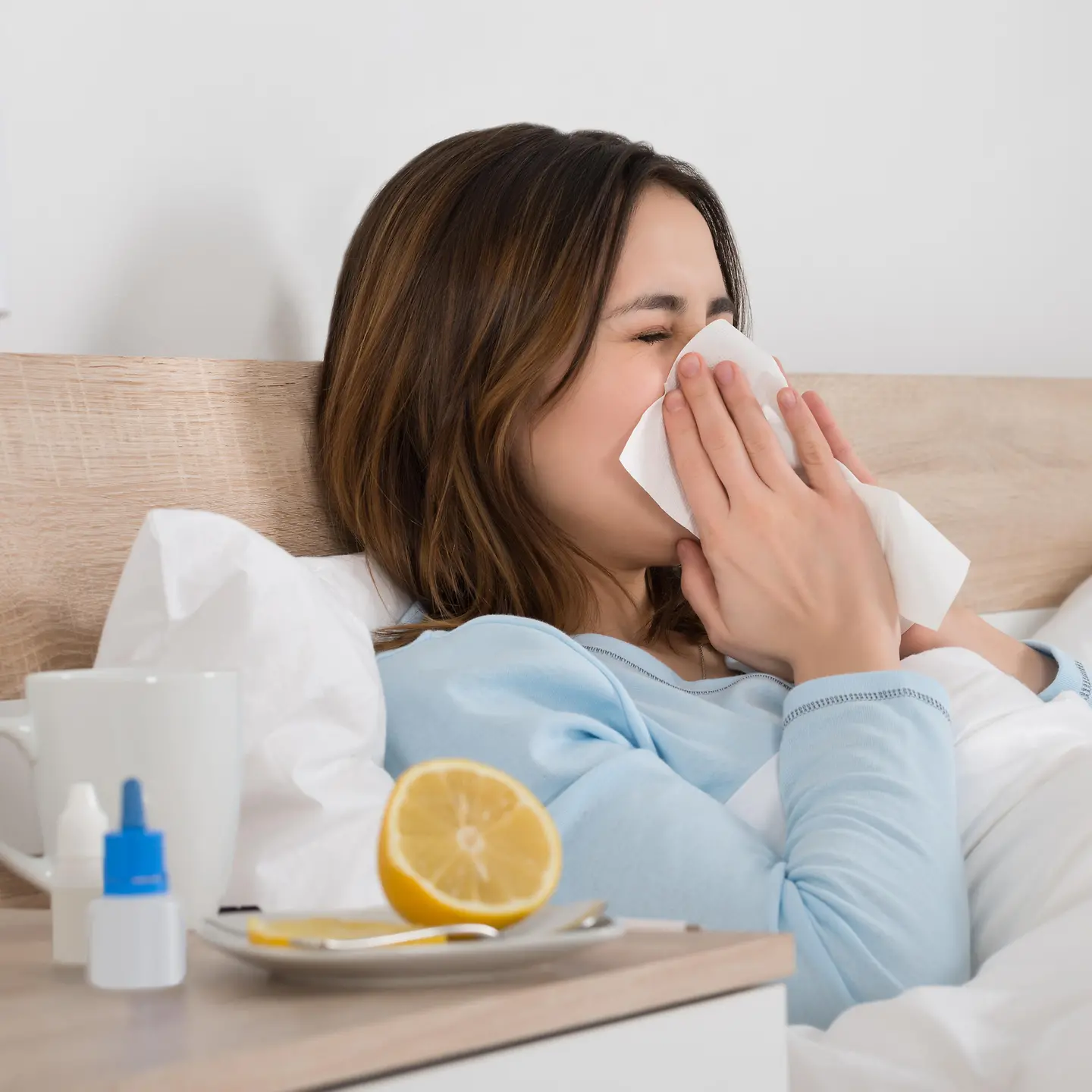 Das Bild zeigt eine Frau, die mit einer Erkältung im Bett liegt und sich die Nase putzt.