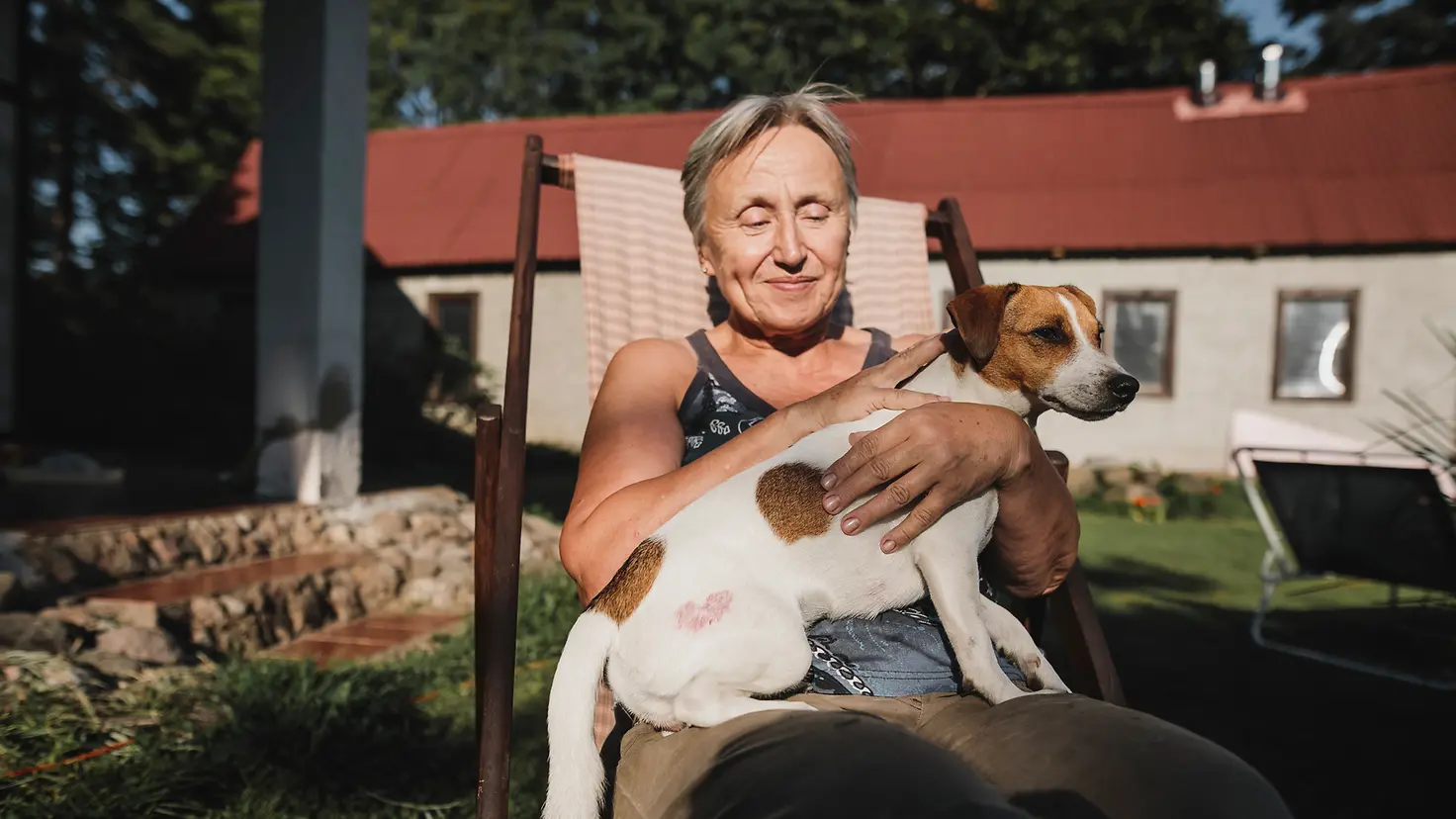 Eine ältere Frau sitzt in ihrem Garten und trägt ihren Hund auf dem Schoss.