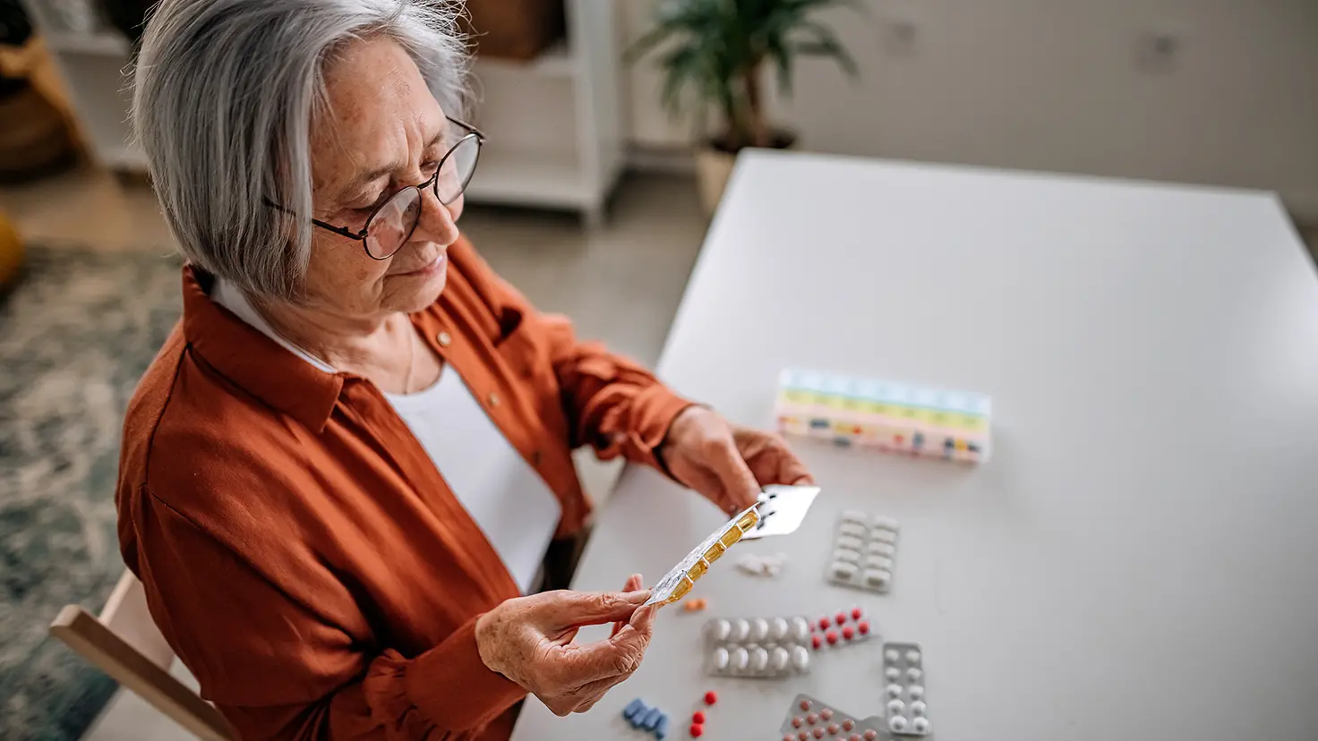 Eine Seniorin sitzt an einem Tisch mit vielen Medikament-Packungen. Sie hält und betrachtet verschiedene Medikamente.