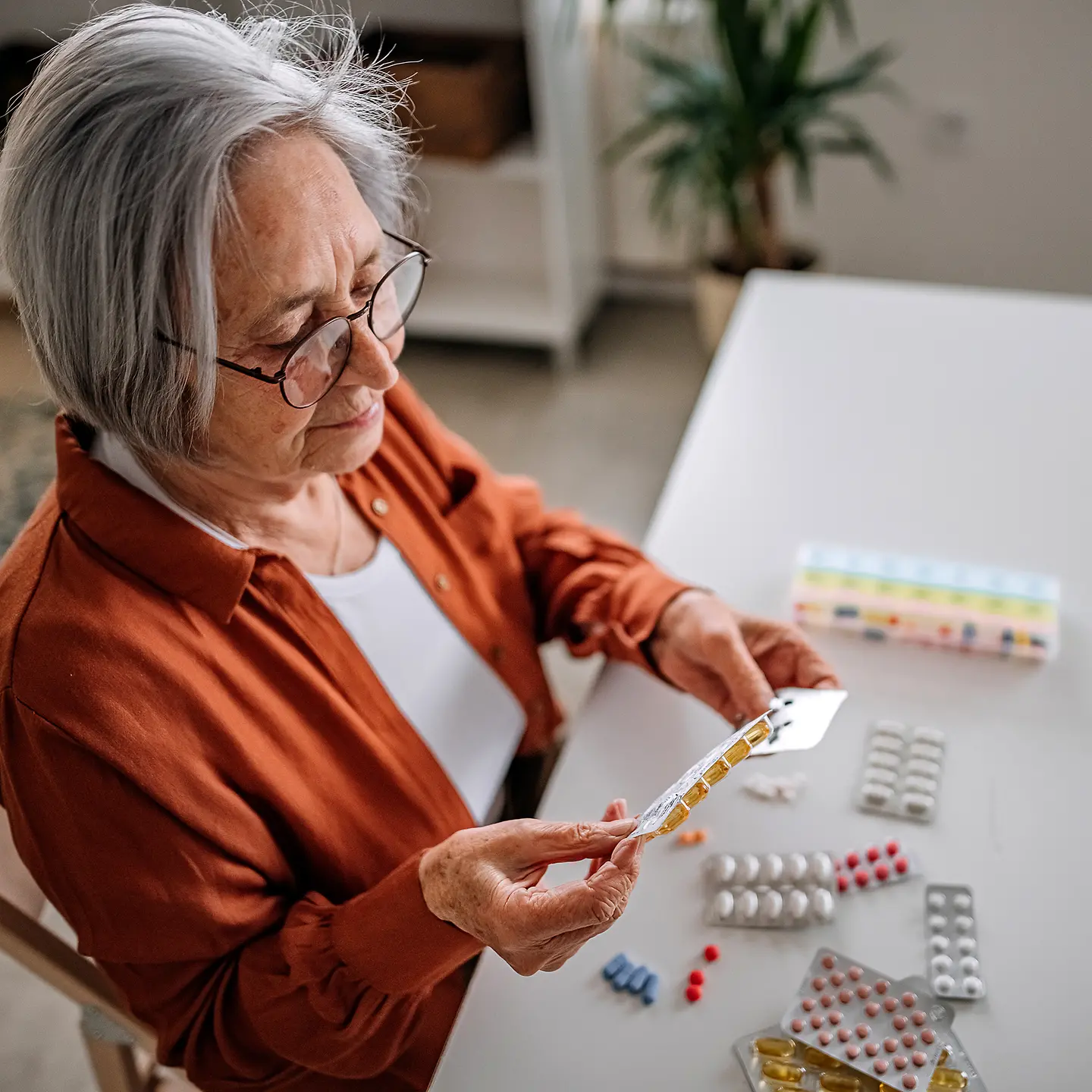 Eine Seniorin sitzt an einem Tisch mit vielen Medikament-Packungen. Sie hält und betrachtet verschiedene Medikamente.