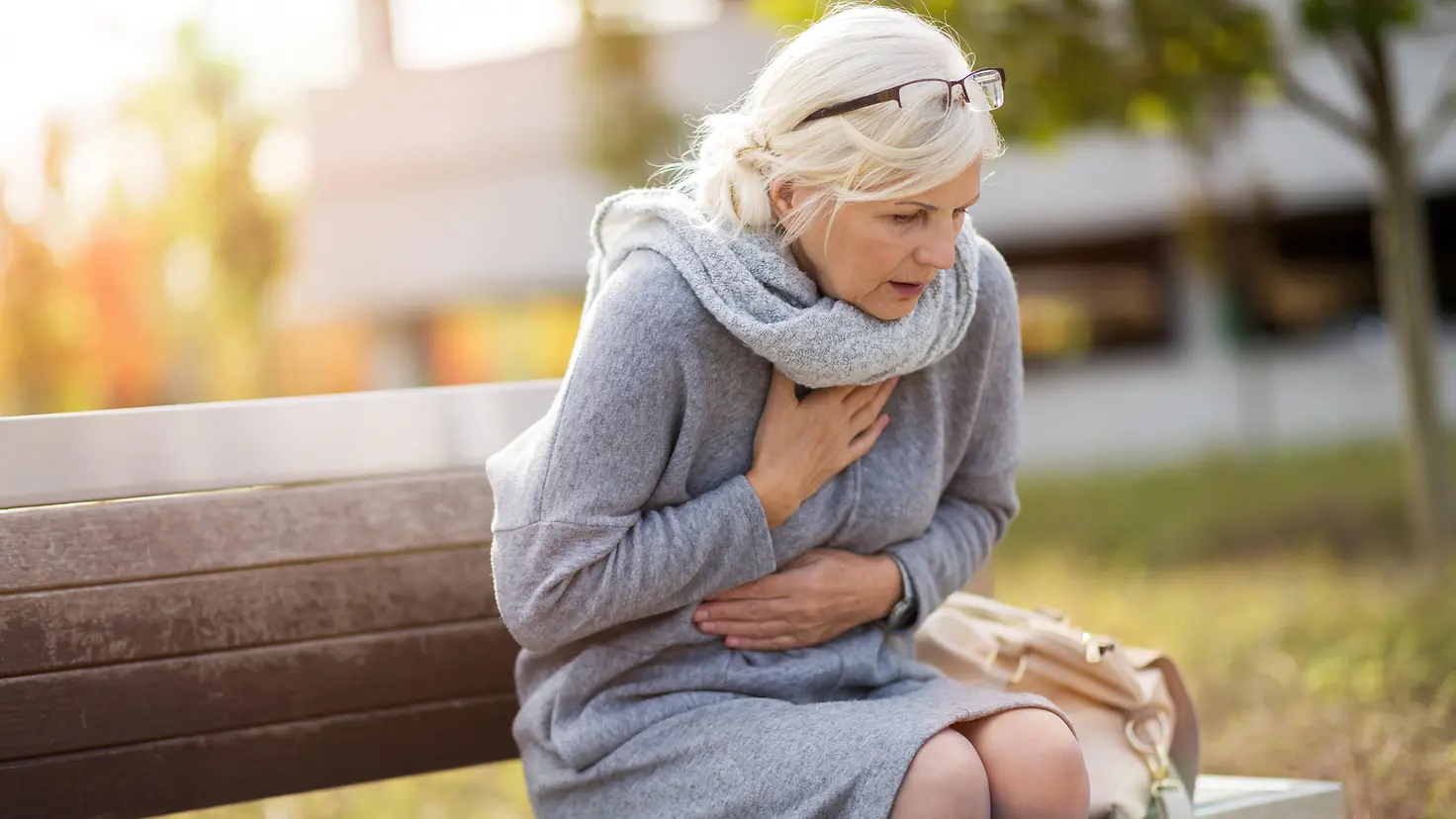 Das Bild zeigt eine Frau, die auf einer Bank sitzt und Symptome eines Herzinfarktes hat.