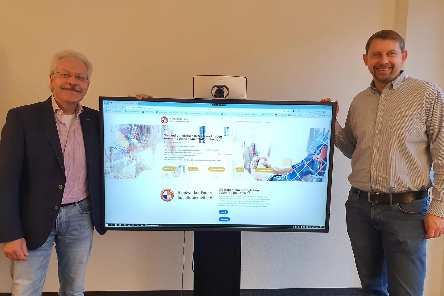 Zwei Männer stehen links und rechts neben einem Präsentationsmonitor, auf dem ein Chart zum Projekt Suchtfrei abgebildet ist 