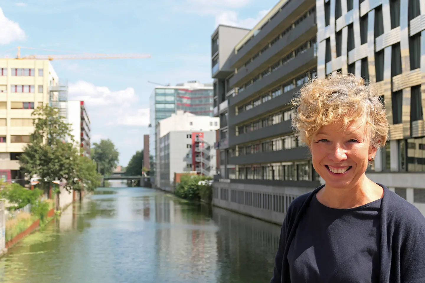 eine lächelnde Frau mit blonden lockigen Haaren steht auf einer Brücke über einen Kanal im Stadtteil Hamburg Hammerbrook