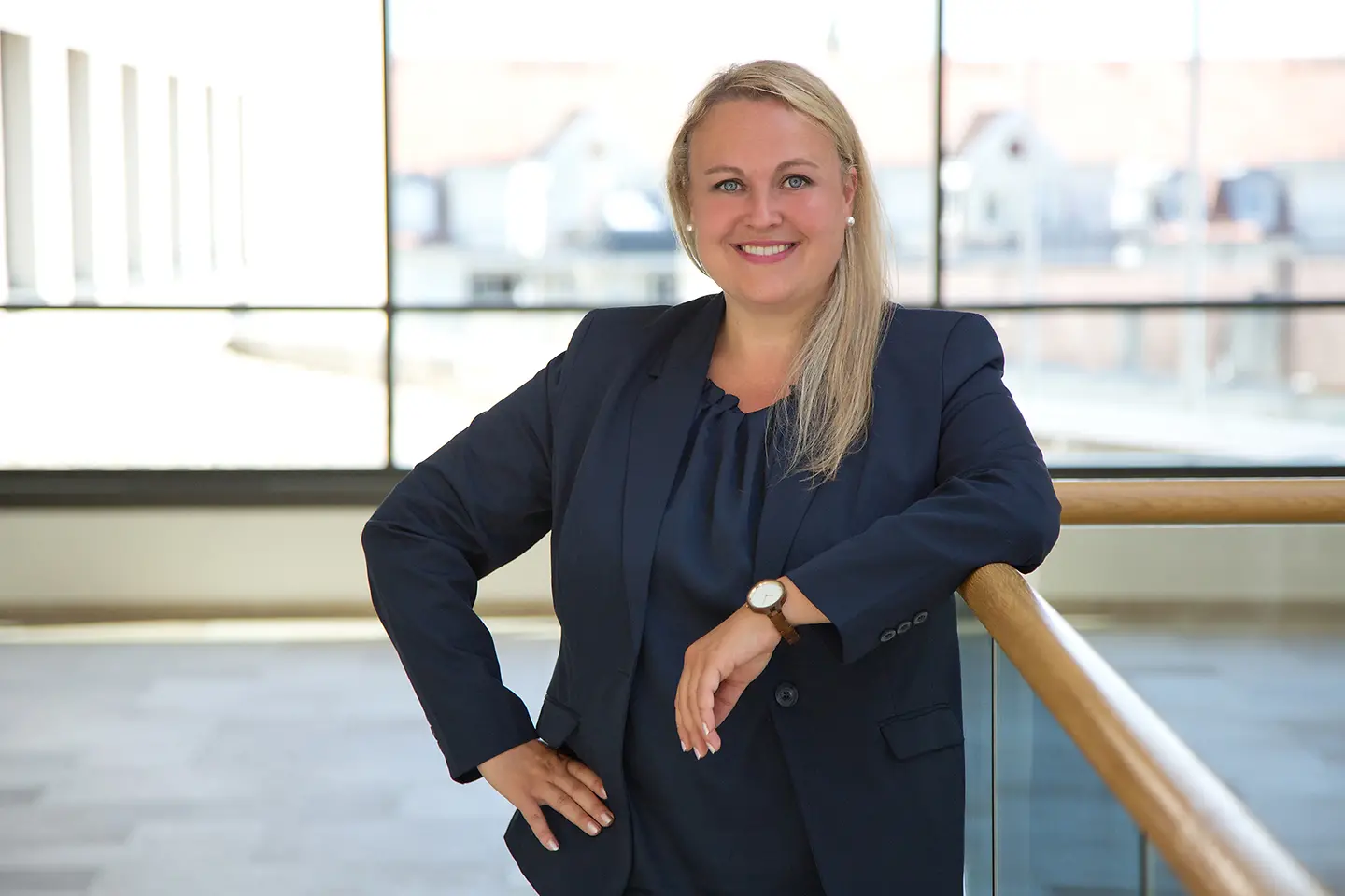Eine junge Frau mit langen blonden Haaren lehnt in einem Bürogebäude an einem Geländer und lächelt in die Kamera. 