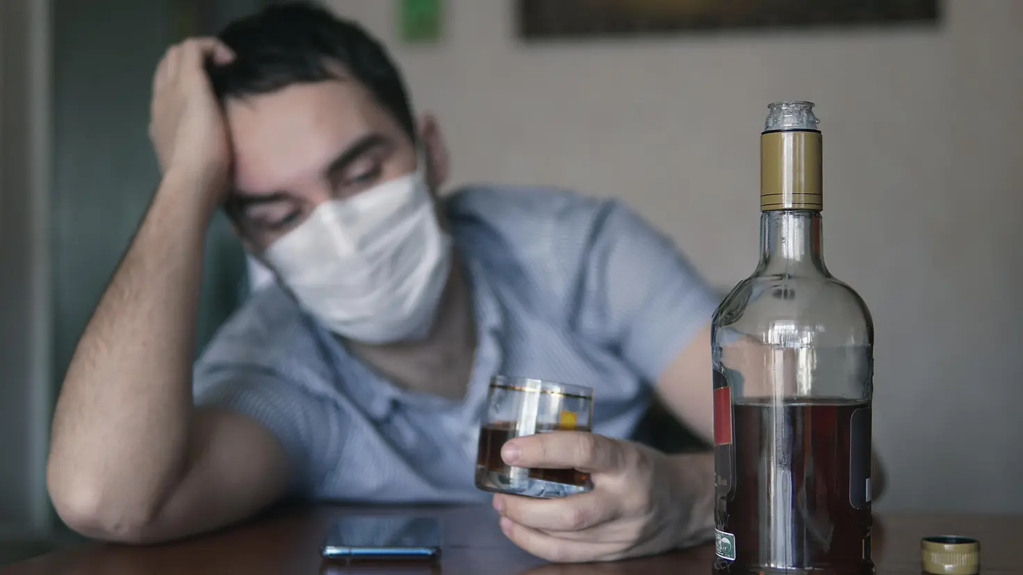 Ein Mann mit Corona-Schutzmaske sitzt an einem Tisch und hält ein Glas mit Alkohol in der Hand. Daneben steht eine Flasche.