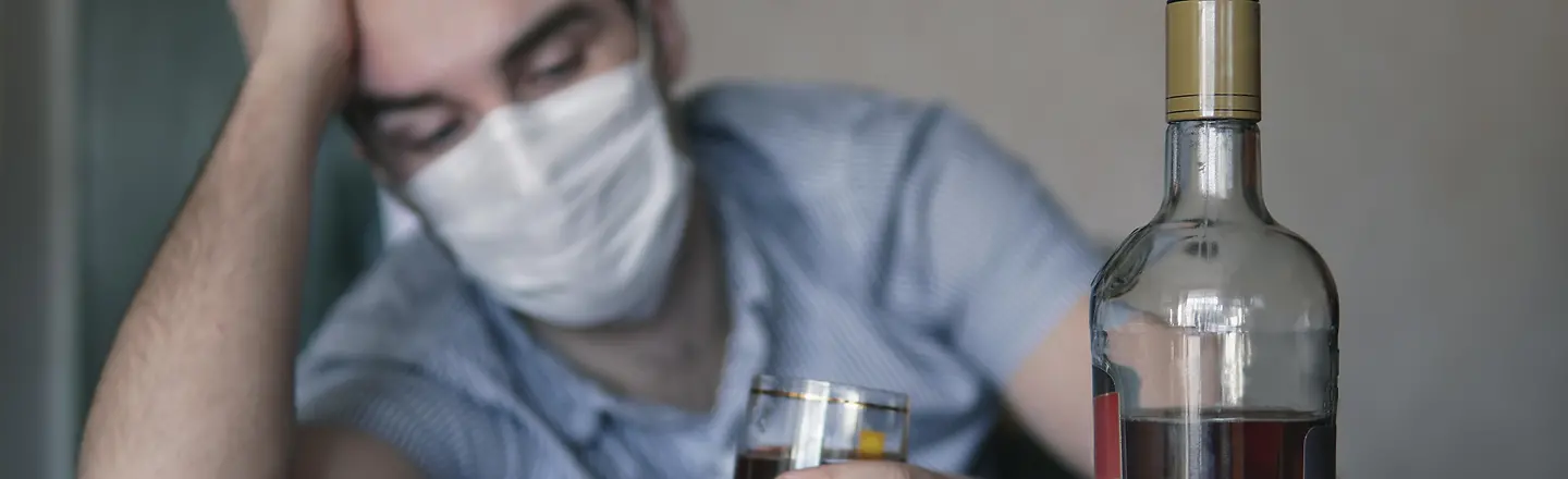 Ein Mann mit Corona-Schutzmaske sitzt an einem Tisch und hält ein Glas mit Alkohol in der Hand. Daneben steht eine Flasche.