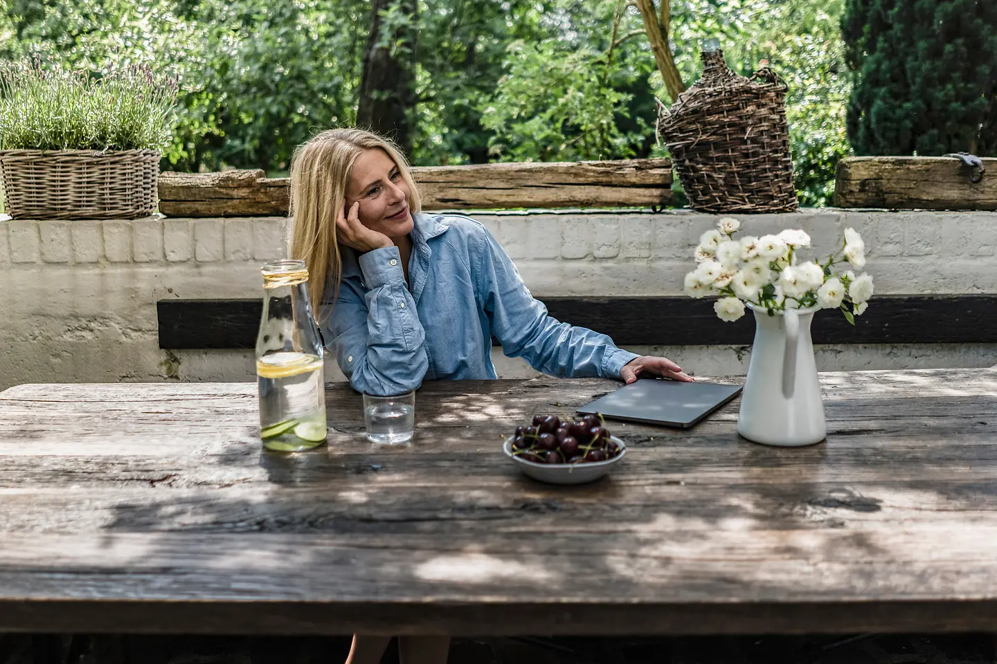Eine Frau sitzt draußen an einem Holztisch auf dem Kirschen, Wasser und eine Vase mit Blumen steht und lächelt in die Luft