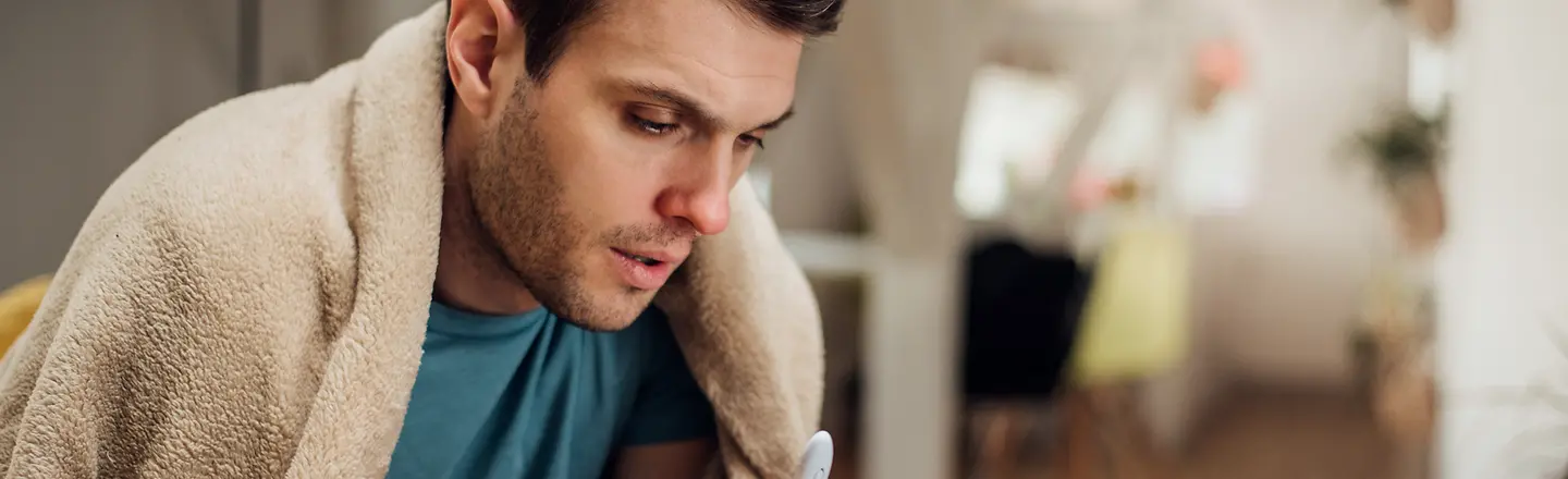 Ein junger Mann mit einer Wolldecke über der Schulter hält ein Fieberthermometer in der Hand und schaut auf ein Tablet