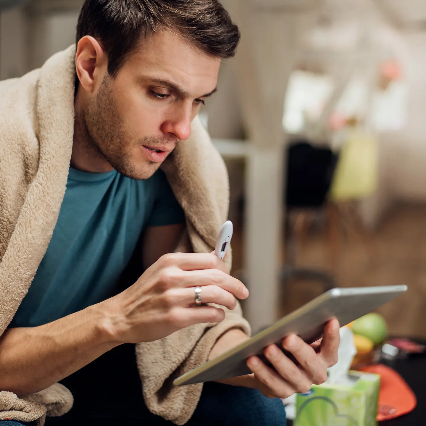 Ein junger Mann mit einer Wolldecke über der Schulter hält ein Fieberthermometer in der Hand und schaut auf ein Tablet