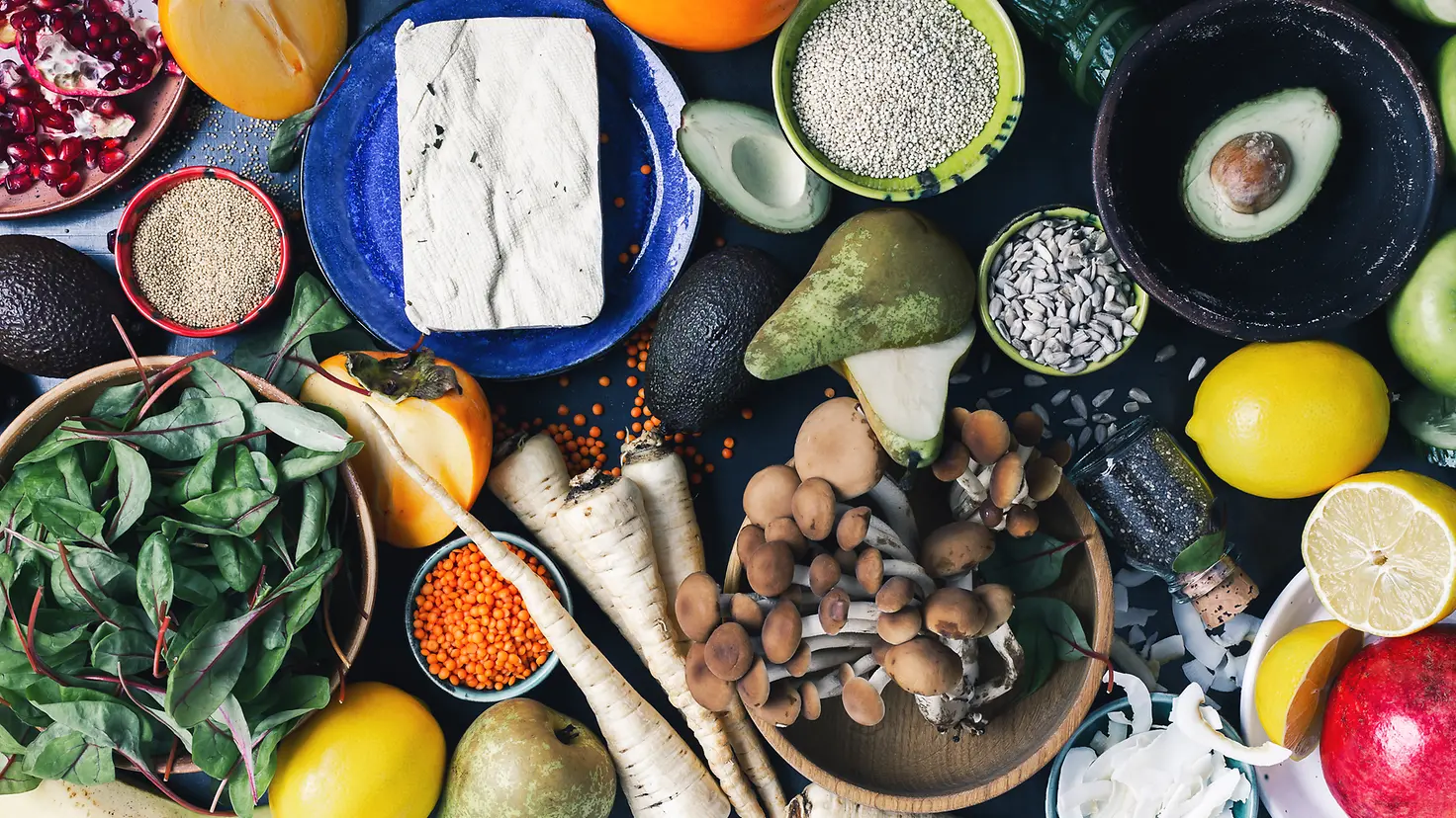 Draufsicht auf einen Tisch mit vielen pflanzlichen Lebensmitteln, frischem Obst, Gemüse, trockenen Körnern und Samen.