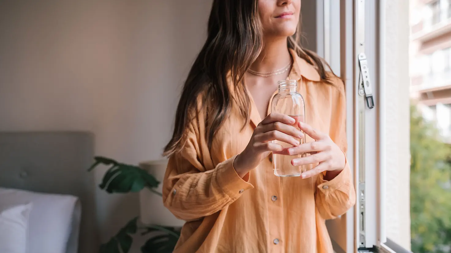 Eine junge Frau mit Wasserflasche blickt aus dem Fenster