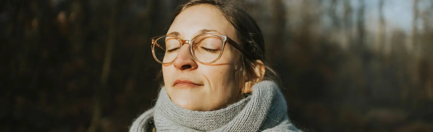 Eine Frau steht im Wald und badet mit geschlossenen Augen in der Sonne