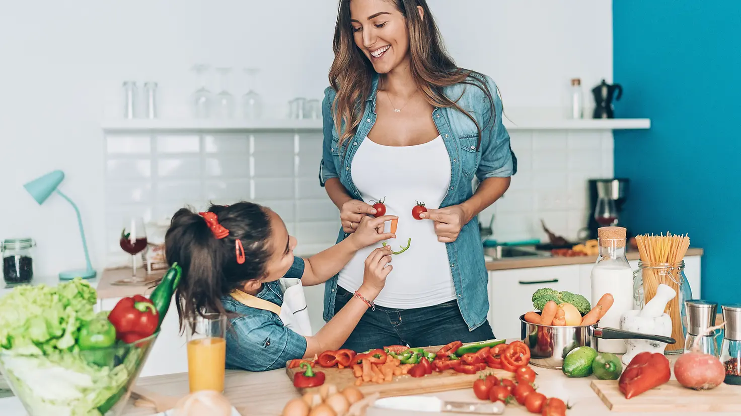 Schwangere Mutter und Tochter machen mit Gemüse ein Smiley Gesicht auf den Bauch der Mutter 