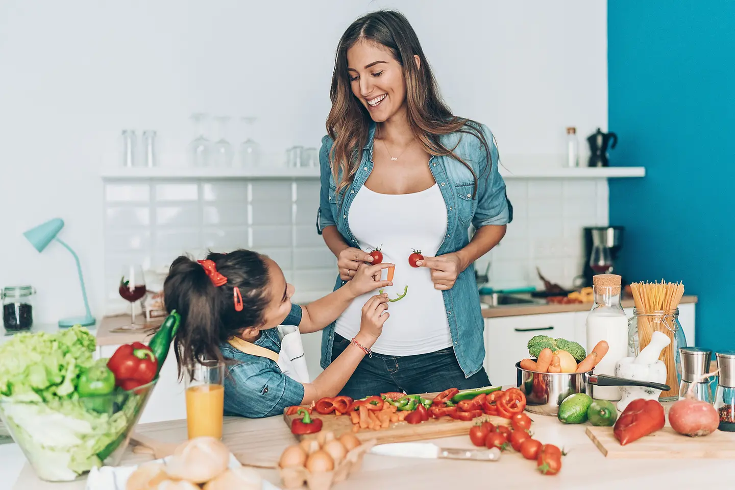 Schwangere Mutter und Tochter machen mit Gemüse ein Smiley Gesicht auf den Bauch der Mutter 