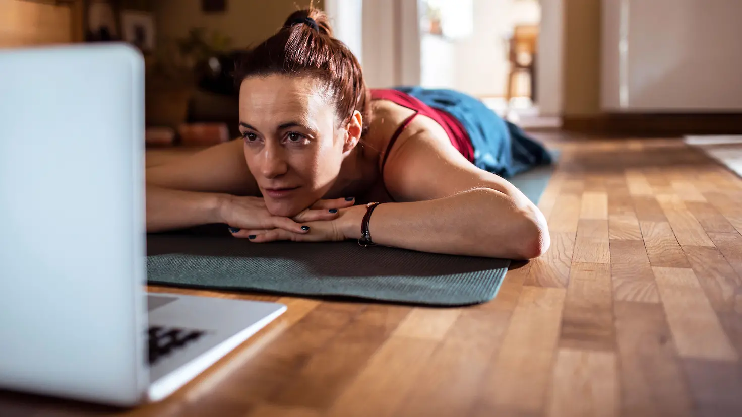 Frau liegt auf einer Yogamatte auf dem Boden und schaut in einen Laptop vor ihr