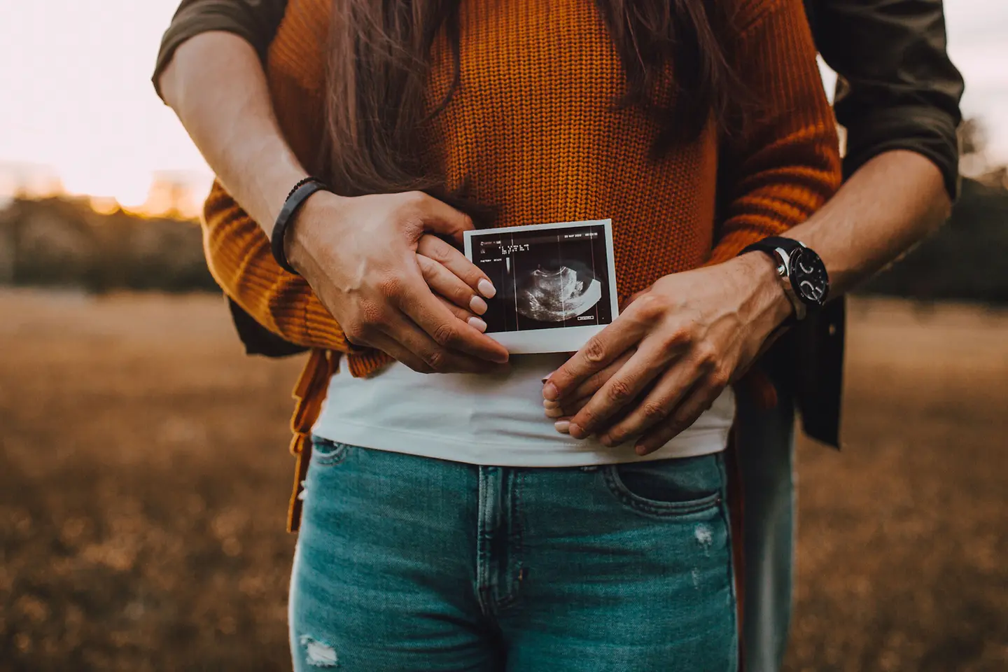 Pärchen steht draußen hintereinander und hält ein Ultraschallbild vor den Bauch der Frau