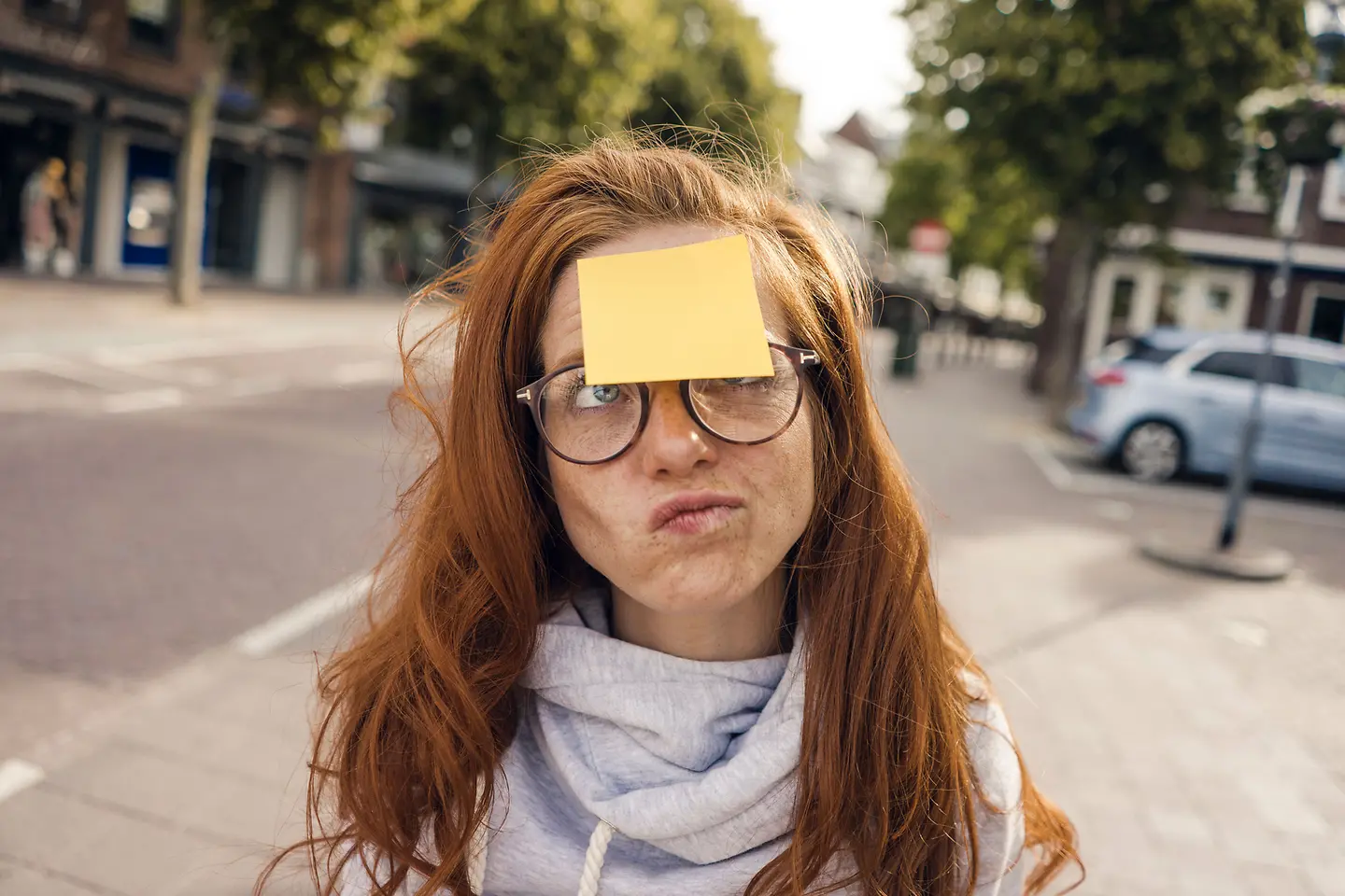 Porträt einer jungen Frau, auf deren Stirn ein Klebezettel klebt und die fragend den Mund verzieht