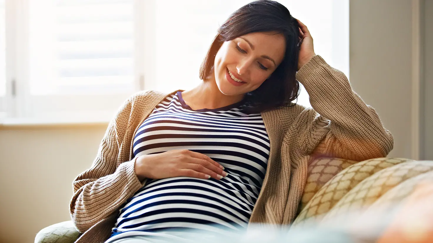Eine Frau in der 17. Schwangerschaftswoche sitzt lächelnd auf einem Sofa legt die Hand auf den Bauch
