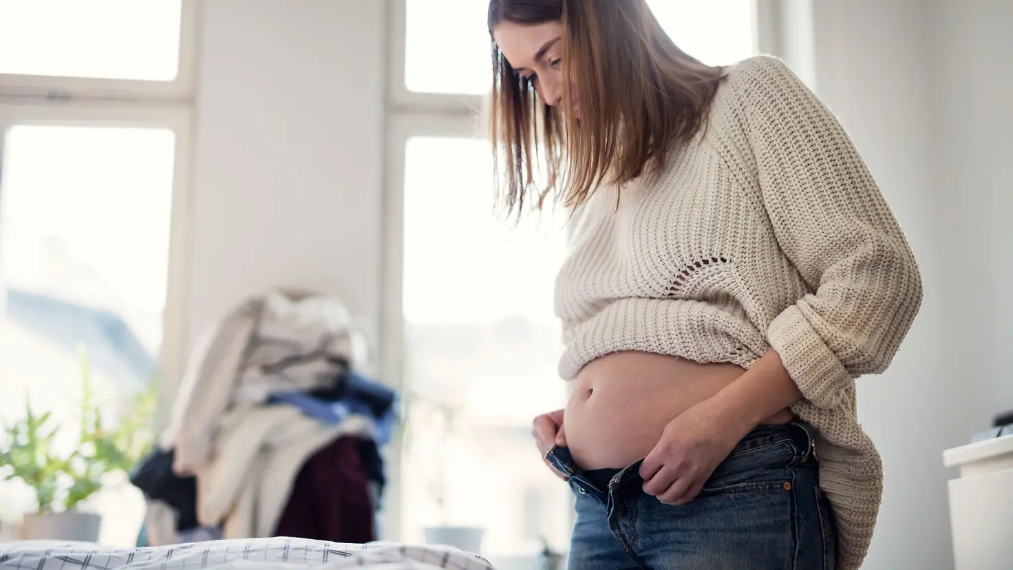 Frau mit Babybauch in der etwa 21. Schwangerschaftswoche versucht Hose zu schließen