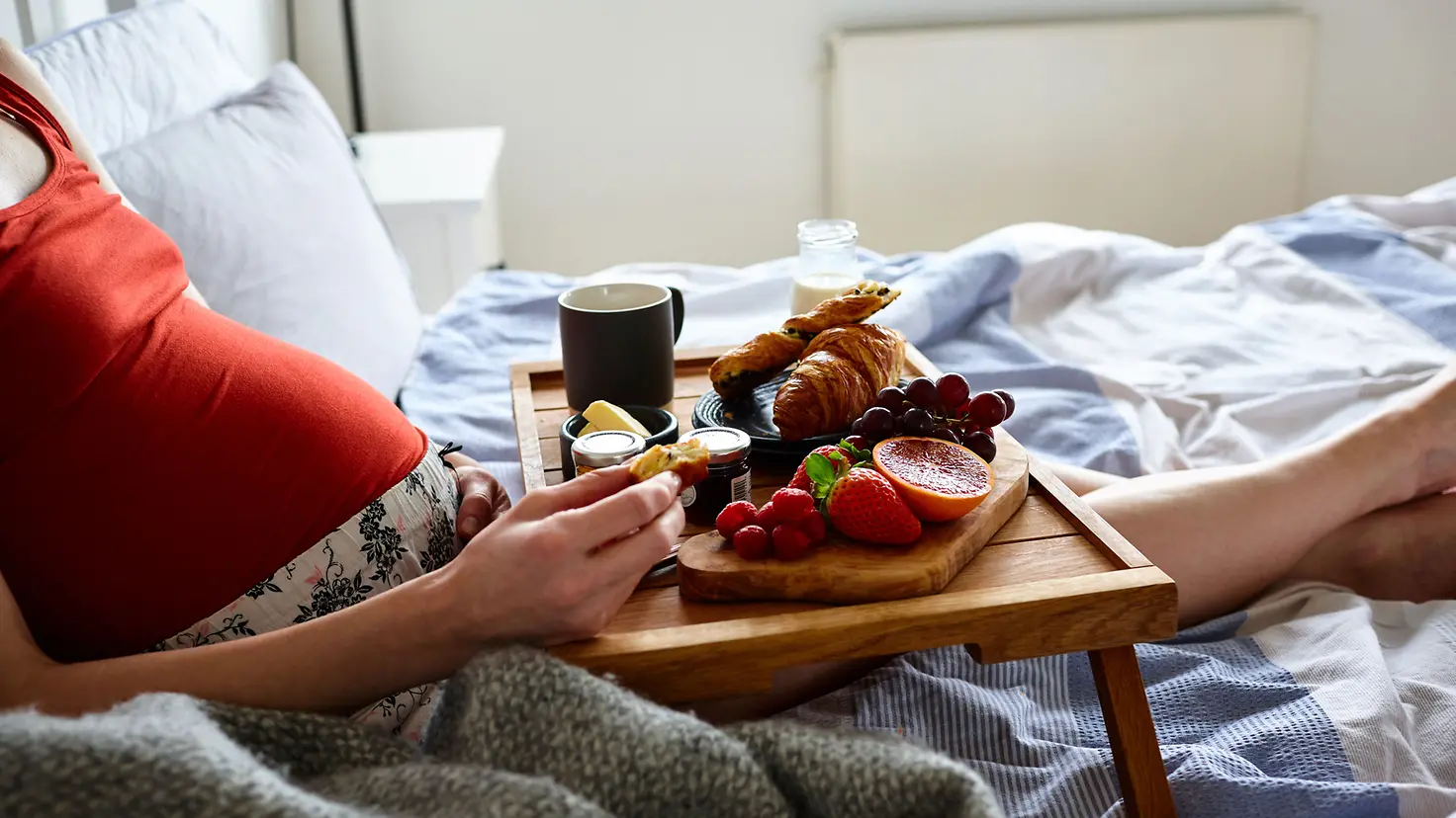 Schwangere Frau in der etwa 22. Schwangerschaftswoche liegt im Bett und Isst Frühstück auf einem Tablett